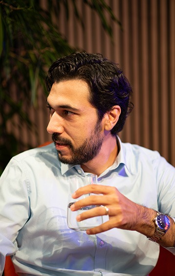 Ein Mann mit dunklem Haar und Bart sitzt in einem Sessel, trägt ein hellblaues Hemd und hält ein Glas Wasser in der Hand.