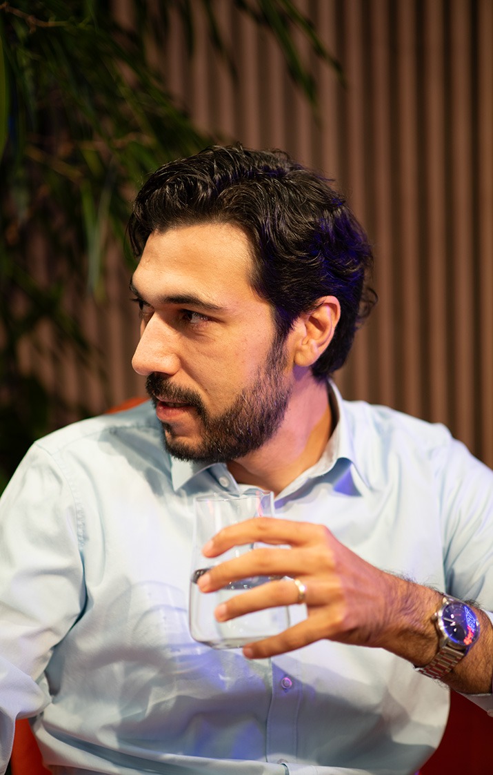 Ein Mann mit dunklem Haar und Bart sitzt in einem Sessel, trägt ein hellblaues Hemd und hält ein Glas Wasser in der Hand.